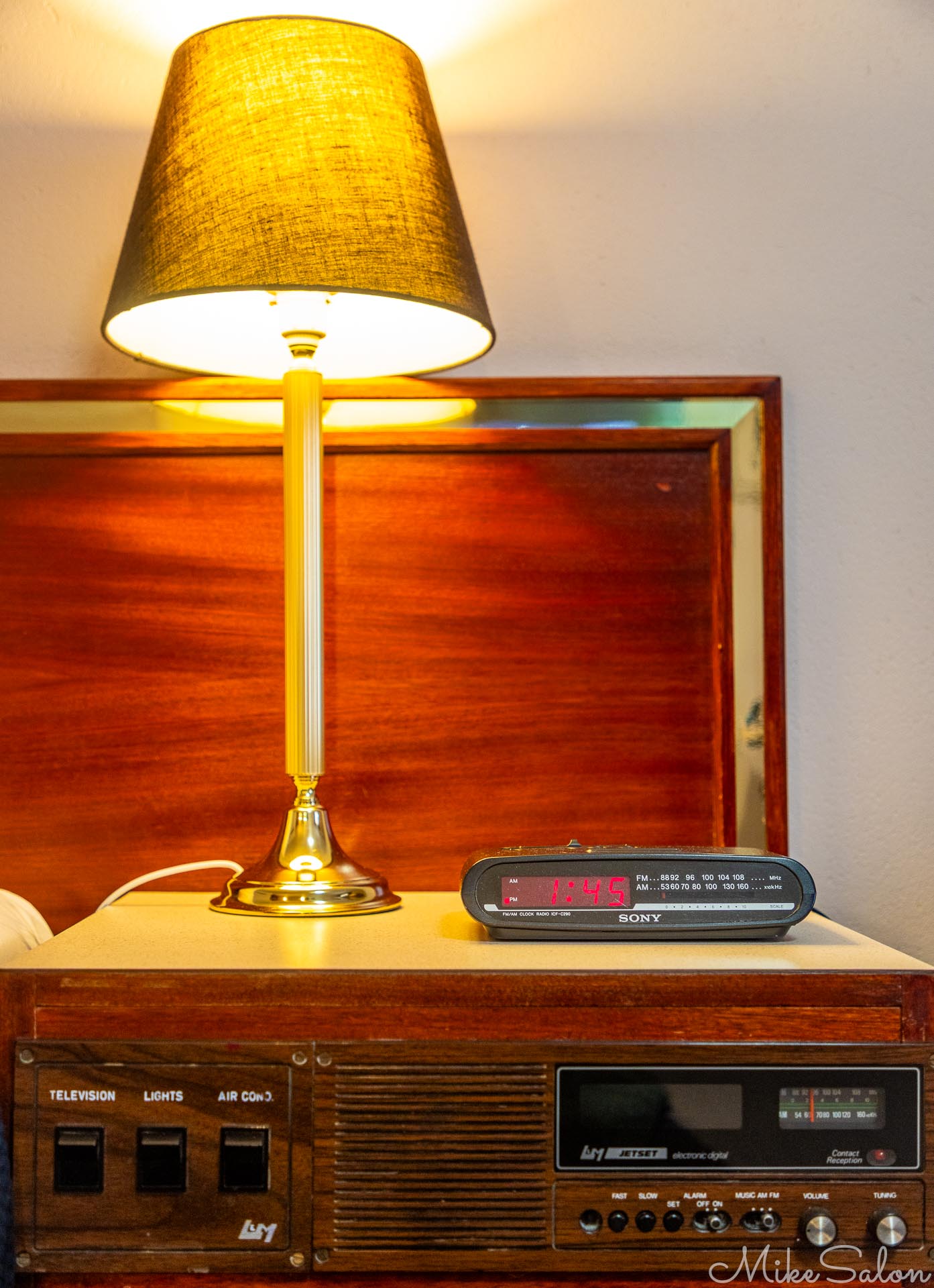 Motel bedside table with lamp, clock radio, and a defunct radio built-in. [5172]