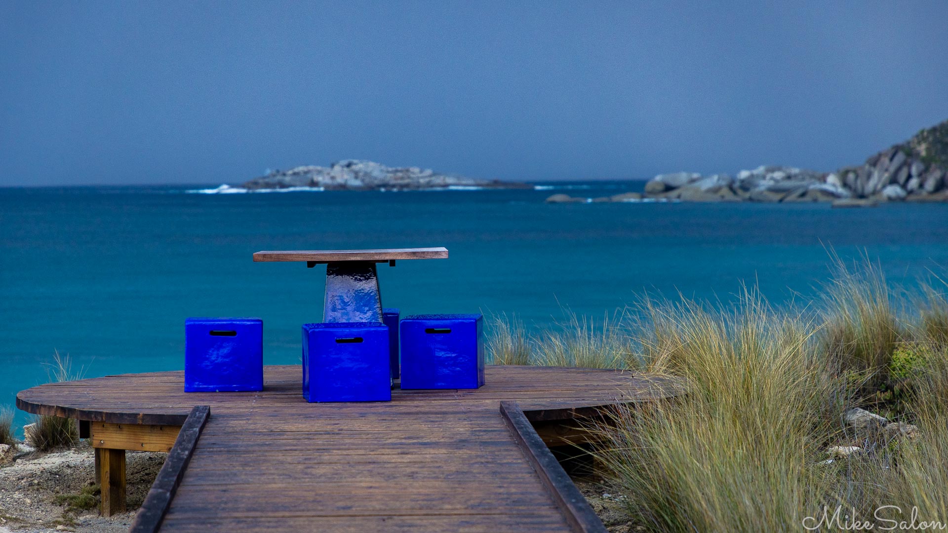 Blue chairs and a picnic table at the end of a boardwalk overlooking the ocean. [4226]