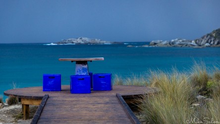 Blue chairs and a picnic table at the end of a boardwalk overlooking the ocean. [4226]