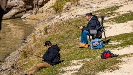 Two fishermen sitting on grassy bank beside river.
