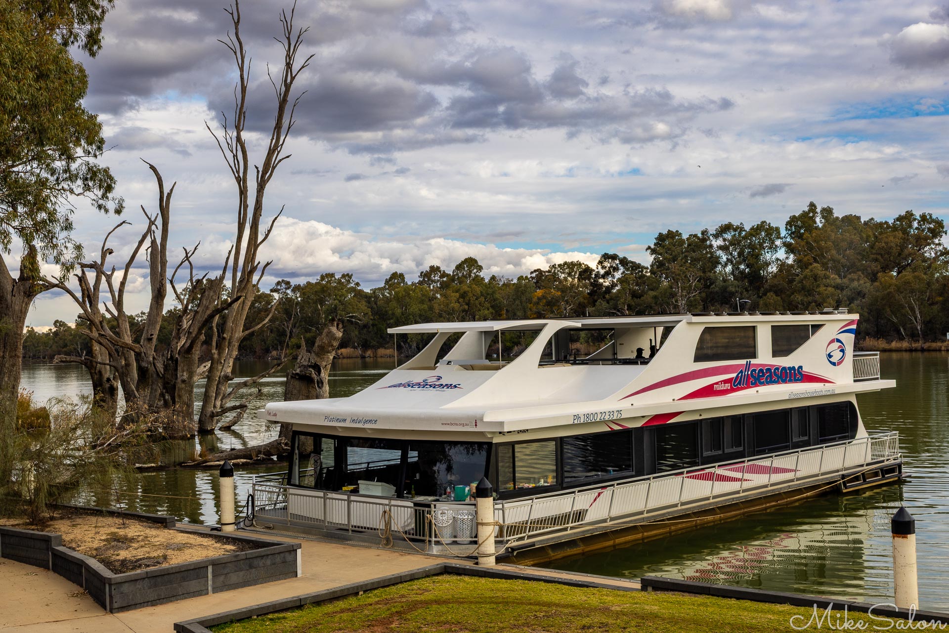 Two‑storey white houseboat moored on calm river with trees in background.