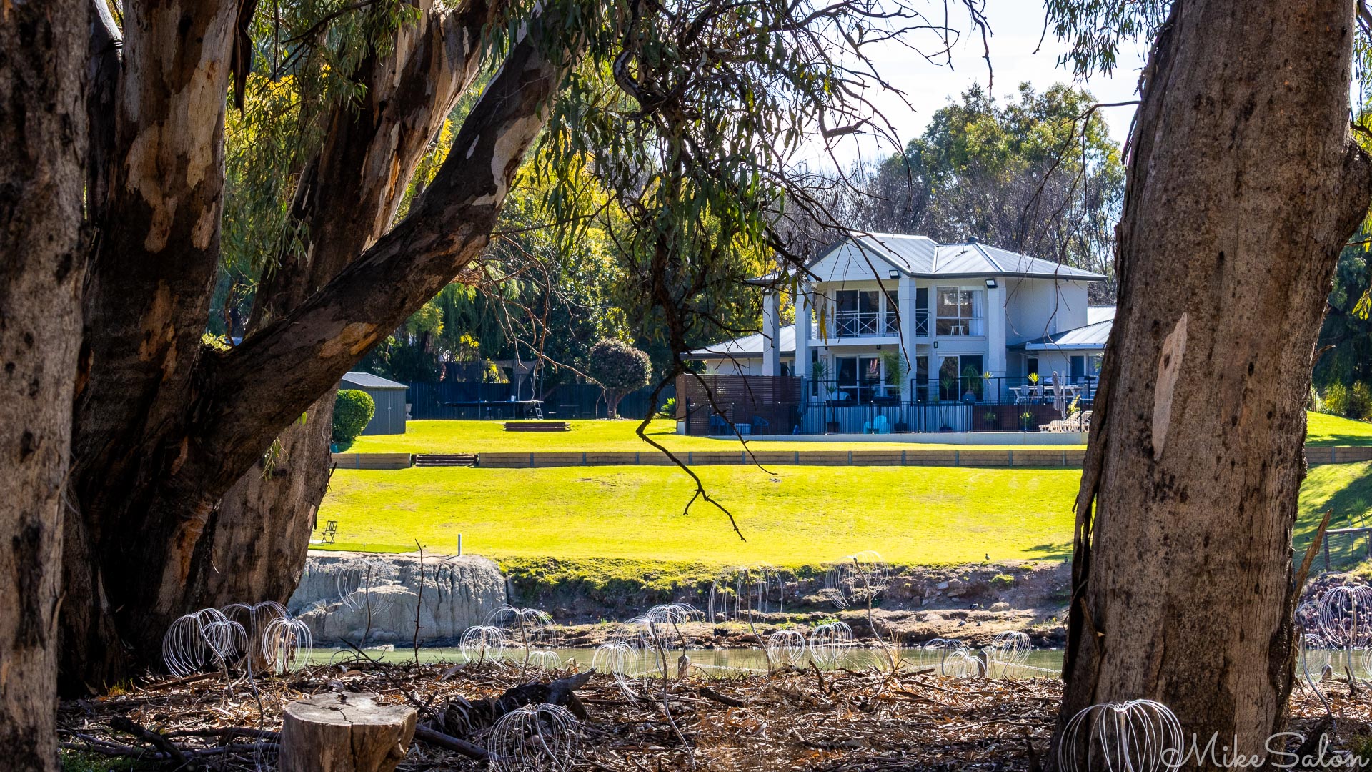 White two‑storey house beside river framed by large gum trees.