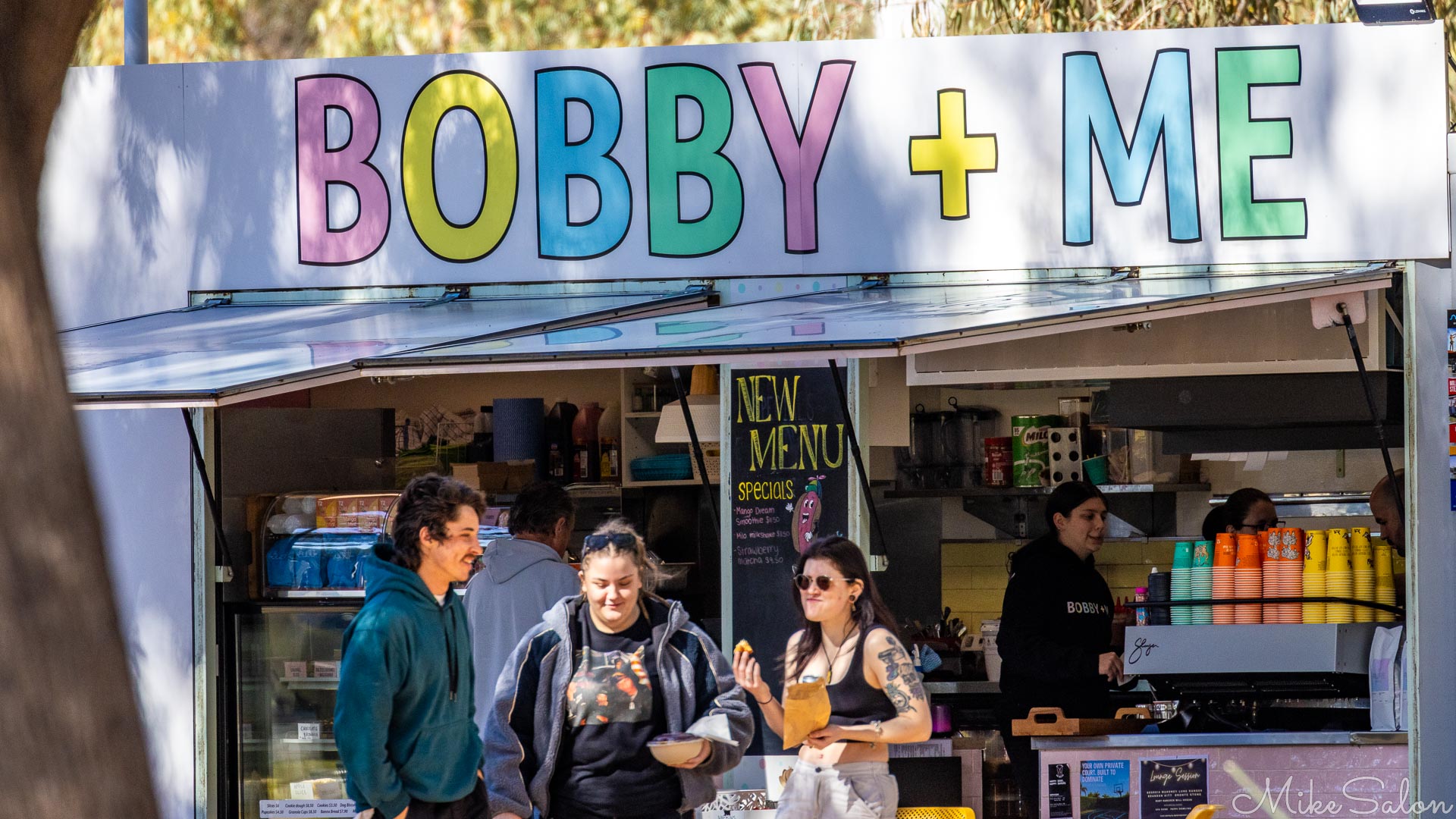 People standing at café counter under sign reading "Bobby + Me."