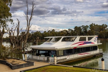 Two‑storey white houseboat moored on calm river with trees in background.