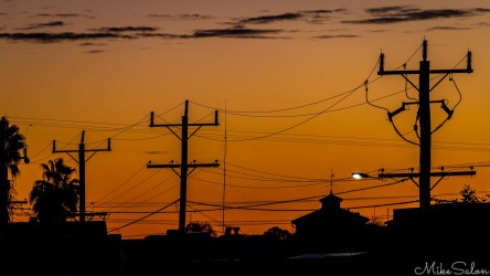 Power poles and cables silhouetted against orange sky at dusk.