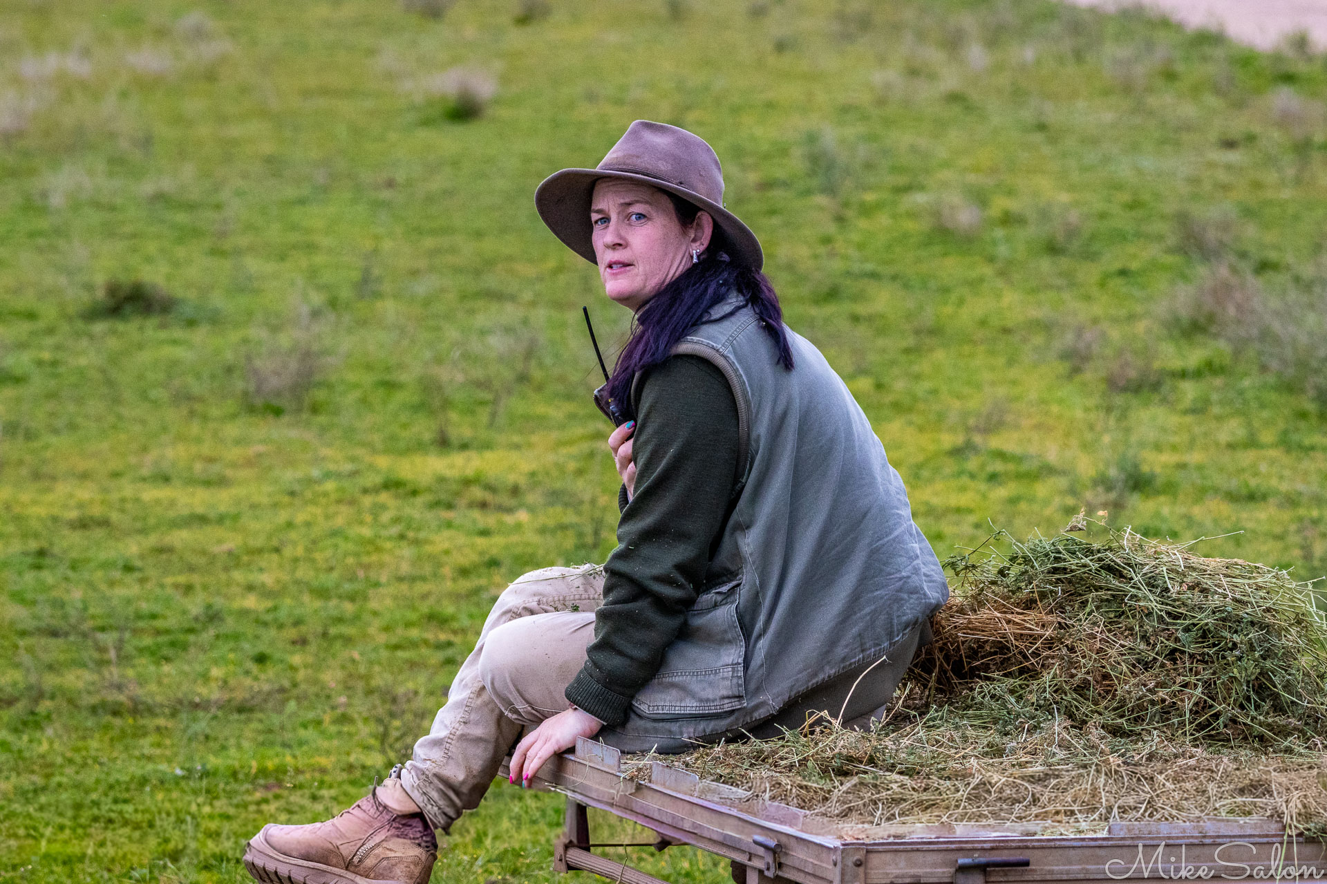 Zoo keeper sitting on a trailer of hay in open grassland, holding a radio while encouraging hippos to approach for feeding. [2547]