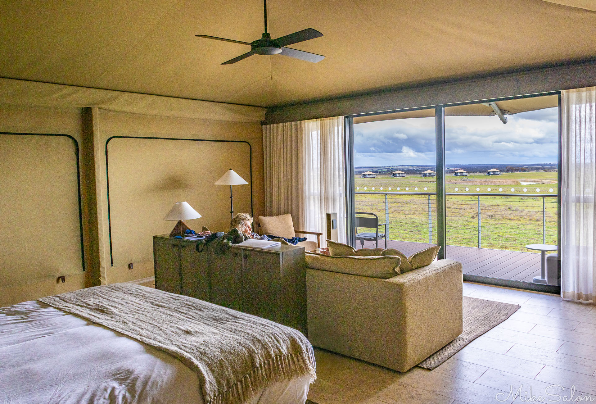 Interior of a luxury safari tent with bed, sofa, and lamps, opening onto a deck overlooking grassy plains and distant lodges under a cloudy sky. [2728]