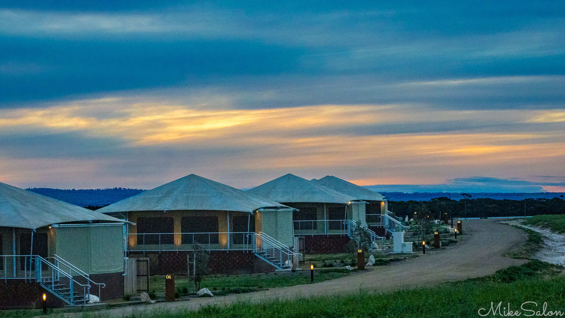 Row of illuminated safari tents along a curved dirt road under a blue‑and‑gold post‑sunset sky, with distant hills on the horizon. [2224]