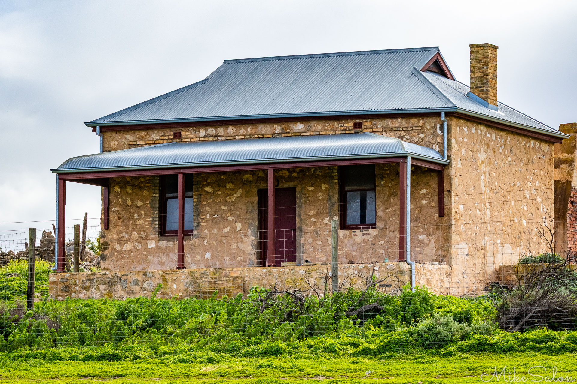Historic stone homestead with corrugated‑iron roof and front verandah, surrounded by green grass and fencing under an overcast sky. [2925]