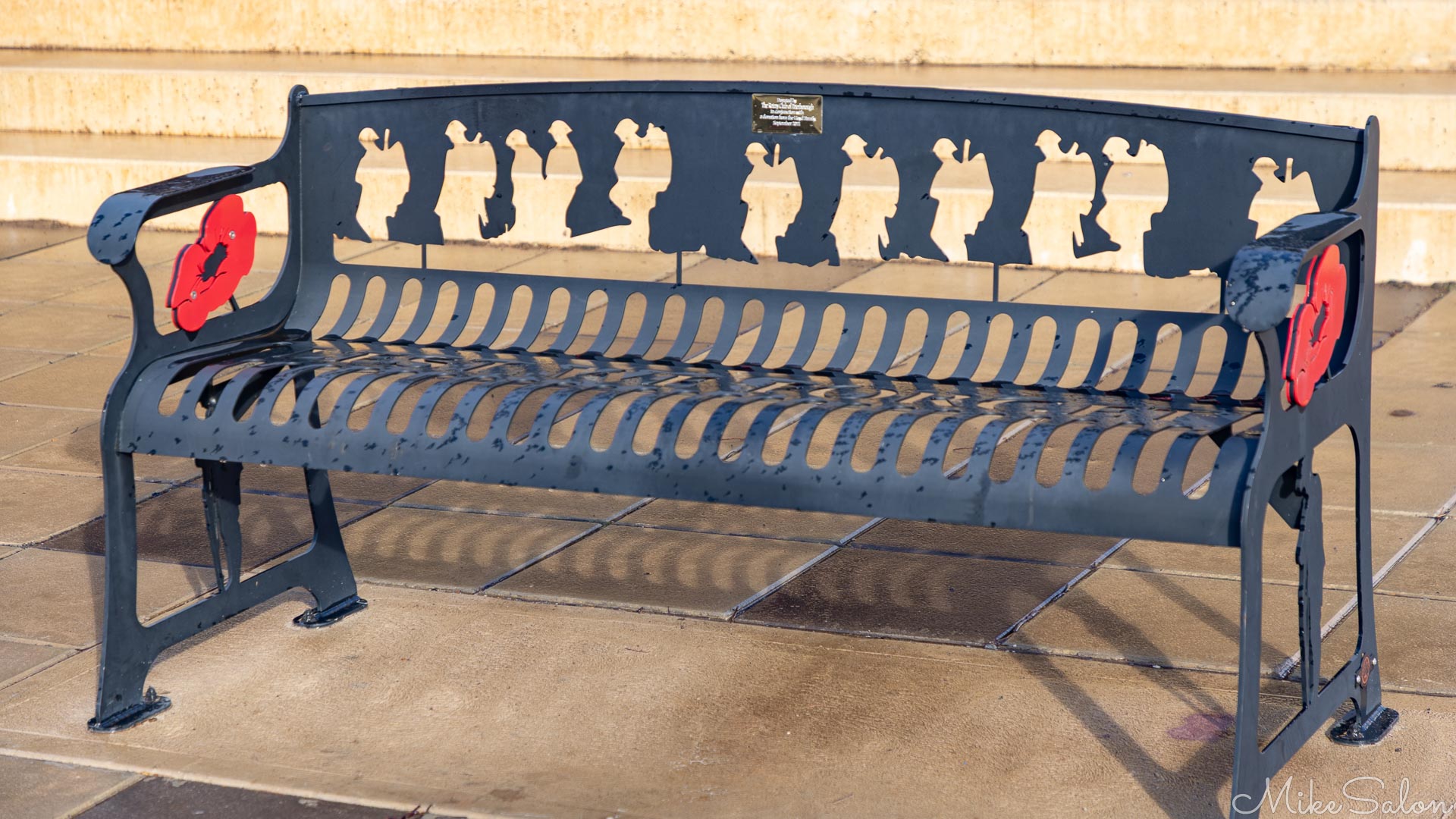 Metal bench with soldier silhouettes and red poppy symbol on paved surface.