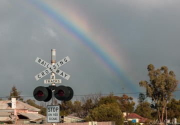 Railway crossing sign and houses under rainbow in cloudy sky.