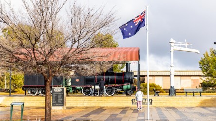 Steam engine under red‑roofed shelter with Australian flag and war memorial area in foreground.
