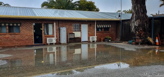Single‑storey brick motel with metal roof and large puddle reflecting building.