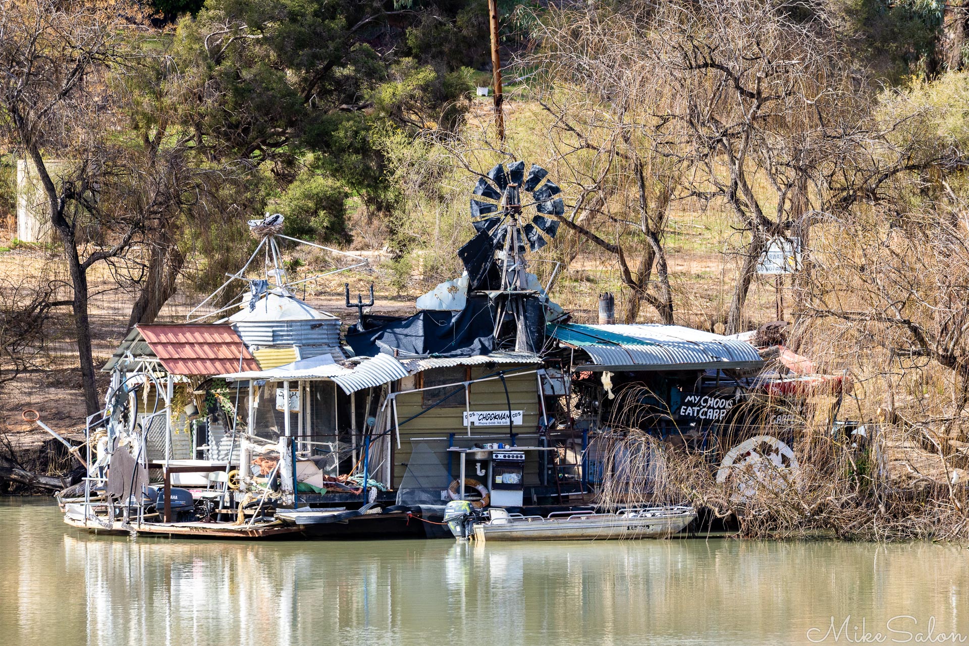 Houseboat‑style structure with windmill and equipment moored on riverbank with trees behind.