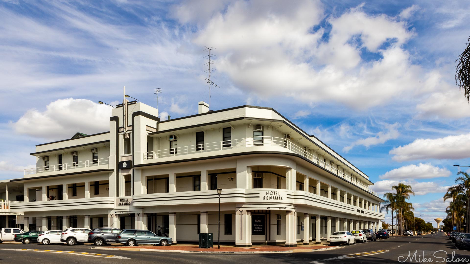 Large three‑storey art deco hotel with white façade, corner balconies, and parked cars under a partly cloudy sky.