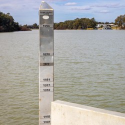 Metal flood marker beside river showing engraved years and water level scale.