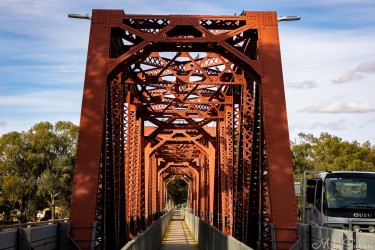 Red steel truss bridge over Murray River with repeating geometric beams and trees in background.