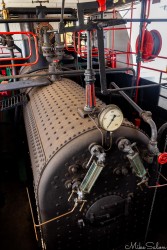 Close‑up of cylindrical steam boiler with pipes, gauges, and red overhead tubing.