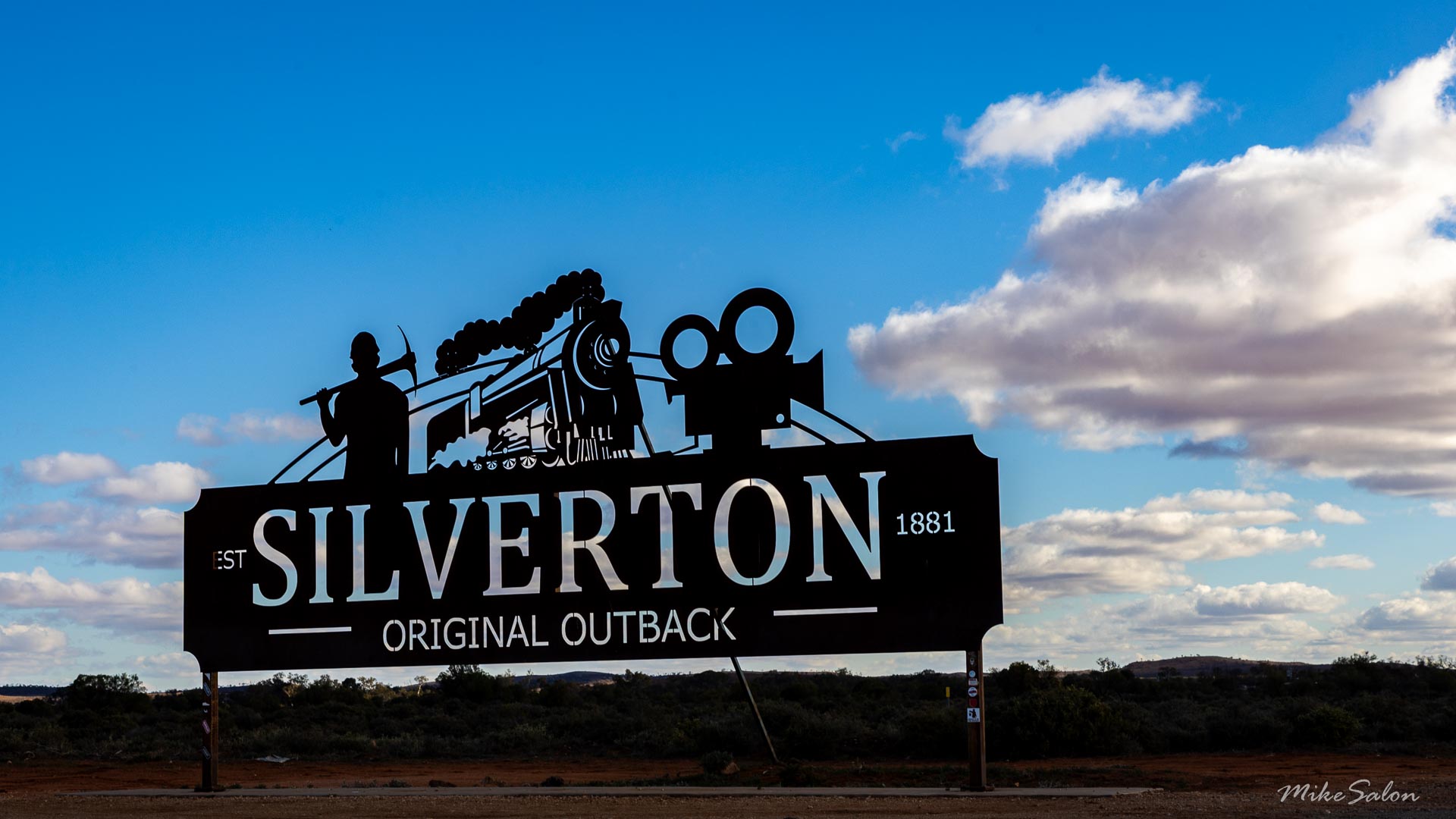 Large roadside sign reading "Silverton Original Outback Est 1881" with silhouettes of train, windmill, and rider against blue sky and desert landscape.