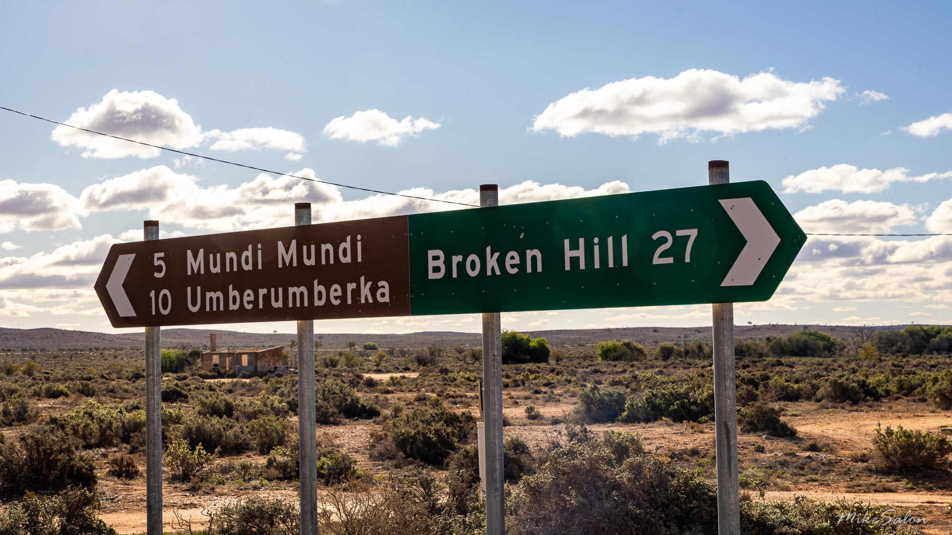 Directional road sign showing distances to Mundi Mundi, Umberumberka, and Broken Hill with scrubland and cloudy sky.