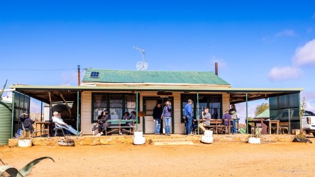 Single‑storey building with shaded porch, tables, and chairs in arid landscape under blue sky.