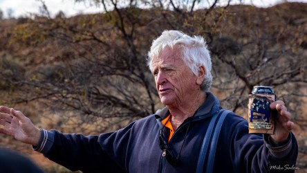 Older man with white hair speaking outdoors, holding a small object, with dry scrubland behind.