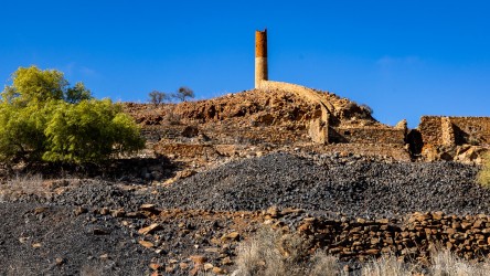  Tall chimney and stone ruins on hill surrounded by rocky terrain under clear blue sky.