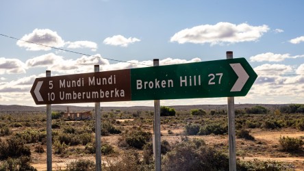 Directional road sign showing distances to Mundi Mundi, Umberumberka, and Broken Hill with scrubland and cloudy sky.