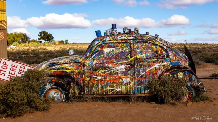 Multicoloured painted VW Beetle surrounded by scrub and sand under blue sky.