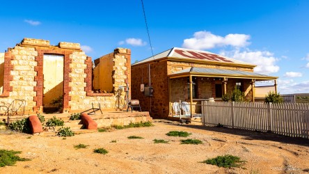 Abandoned cafe on right, with stone ruins with partial walls on the right and sandy ground under bright blue sky.