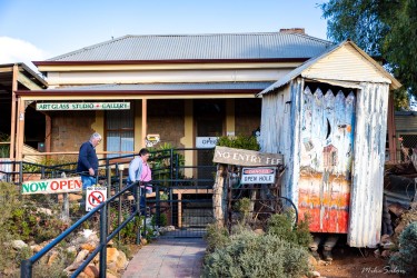 Small corrugated‑iron building with porch, signs, and artworks outside, including "Now Open" notice, and a pretend occupied outdoors toilet out the front.