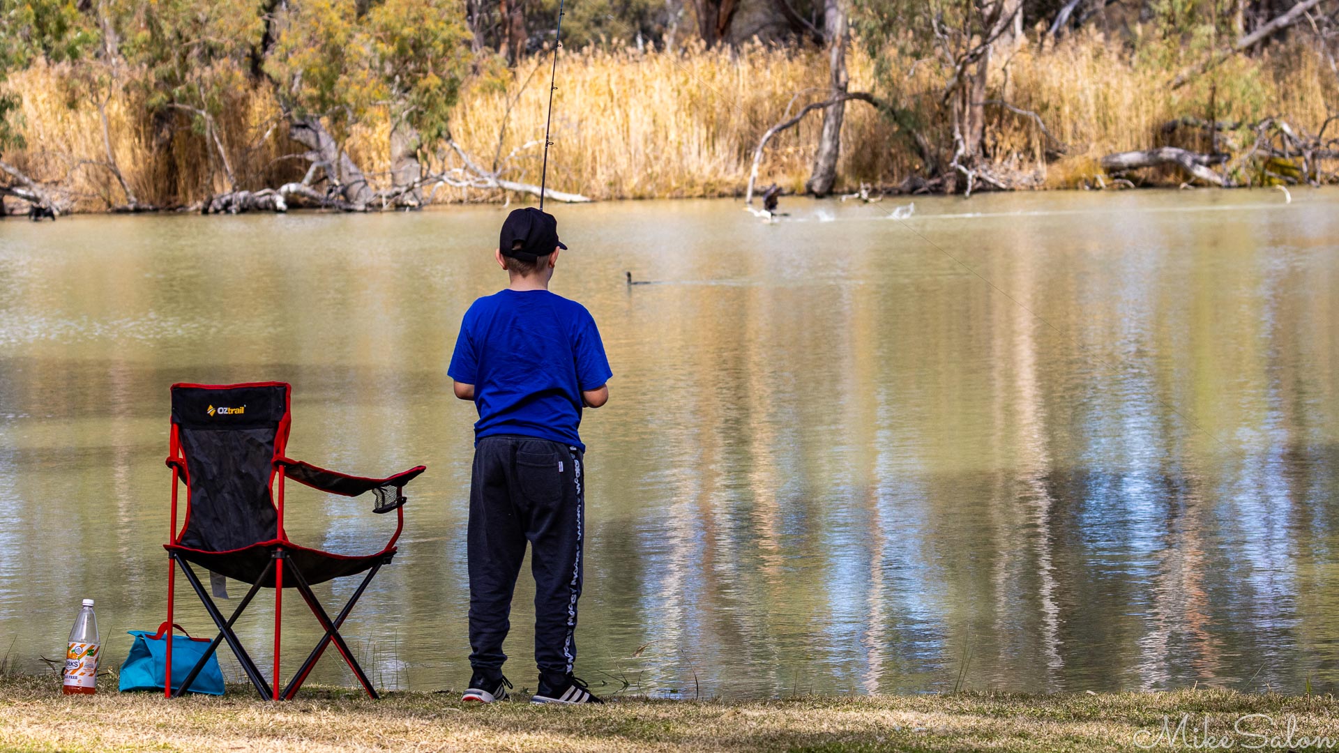 Young person standing beside chair and fishing gear on riverbank with calm water and trees.