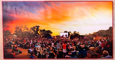 Large crowd gathered outdoors at sunset with stage and glowing orange sky.