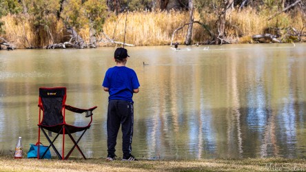 Young person standing beside chair and fishing gear on riverbank with calm water and trees.