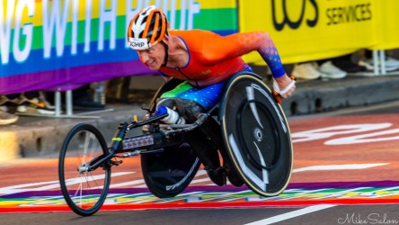 Wheelchair racer in bright orange helmet and multicoloured suit pushing hard, with rainbow banners and spectators in the background.