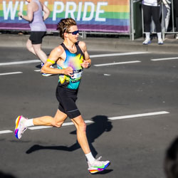 At the 32 km mark, a runner powers through Taylor Square in perfect form, eyes fixed ahead and mind locked on the finish. The “Running with Pride” banner behind him mirrors his own vibrancy and determination. [3890]
