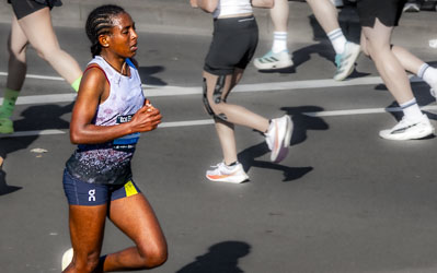 One of the leading female runners powers through Taylor Square at the 32 km mark, her concentration absolute as she glides past the outbound crowd on the opposite side of Oxford Street. The sunlight catches her form, highlighting the calm determination of an elite athlete deep in the race. [3891]