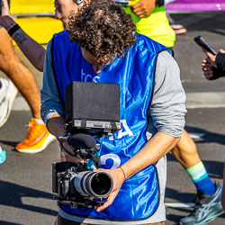A media crew member films the action at Taylor Square, focusing on the surge of runners and spectators along Oxford Street. His concentration mirrors the athletes’ own intensity. [3915]