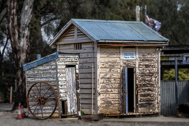 Weathered timber police cells from circa 1880 at Tatura, with corrugated iron roof, wagon wheel, and Australian flag in background.