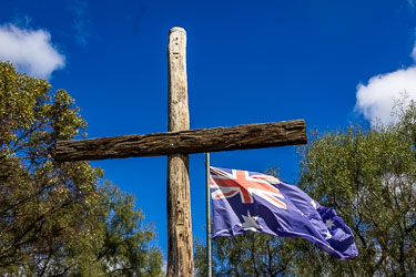 Wooden memorial cross with Australian flag flying beside it under a bright blue sky, commemorating servicemen and veterans at the Great Aussie Beer Shed.