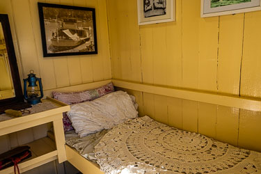 Small recreated cabin aboard the P.S. Pevensey with yellow wooden walls, lace bedspread, pillows, lantern, and framed riverboat photographs.