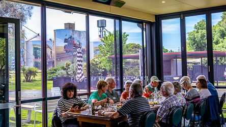 Group of Australian Star passengers dining at a long table inside the Colbinabbin Hotel, with large windows showing silo art of a steam train and rural scenery outside.