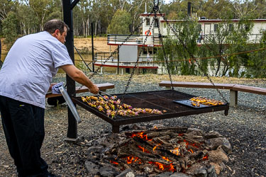 Chef tending skewers on a suspended woodfire barbecue near the Murray River with the Australian Star moored in the background.
