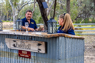 Paul and Julie from the Australian Star at an outdoor corrugated‑iron bar with P.S. Emmylou signage and a 2022 Flood Level sign, surrounded by river gums at Layfield Lane.