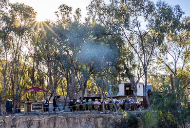 Passengers seated at long tables under tall river gums at Layfield Park, enjoying a barbecue dinner beside the Murray River in warm evening light.