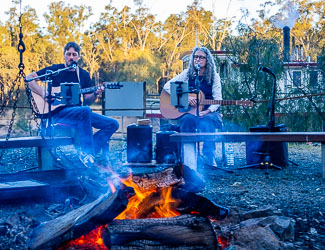 Two guitarists performing beside a campfire at Layfield Lane with a paddle steamer moored in the background, evening light filtering through river gums.