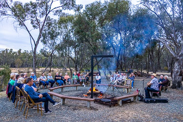 Musician Warren performing with guitar beside a campfire at Layfield Lane, surrounded by seated guests under tall river gums near the Murray River.