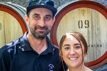 Portrait of Drew and Sarah, owners of Murray Gums Estate, standing before wine barrels in Moama.