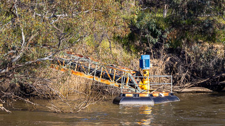 Floating mechanical water pump with a blue motor and metal intake arm extending from the riverbank into the Murray River, surrounded by reeds and gum trees.