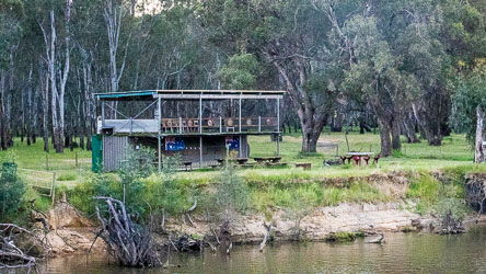 Simple riverside shed made of corrugated metal with an open deck and chairs overlooking the Murray River, surrounded by gum trees and grassy floodplain.